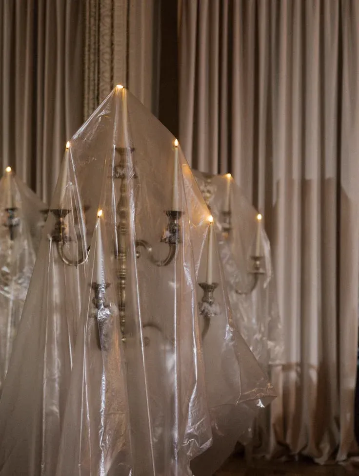 Chandeliers recouverts d'un voile plastique transparent, formant des silhouettes de fantômes éclairées par des bougies, devant un rideau beige