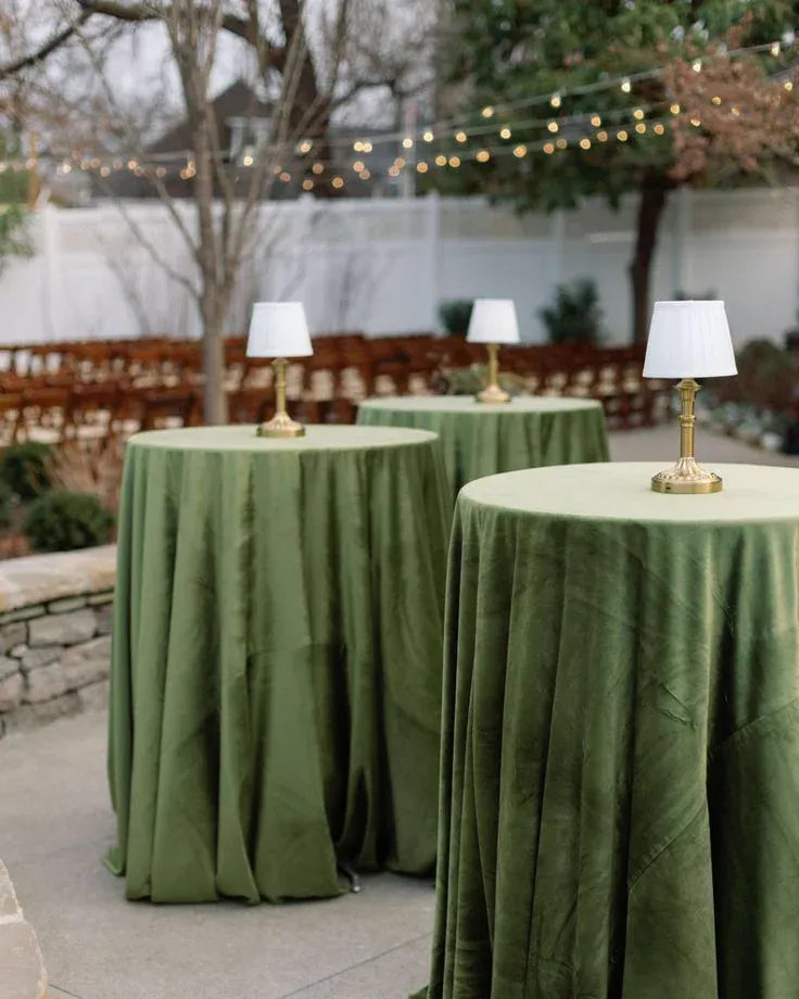 Trois mange-debout recouverts d'une nappe de velours vert et surmontés de petites lampes, dans un jardin éclairé par une guirlande lumineuse
