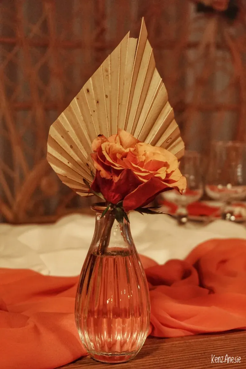Vase en verre strié à bord doré portant une rose orangée et une palme séchée, sur une table dressée avec un voile orange